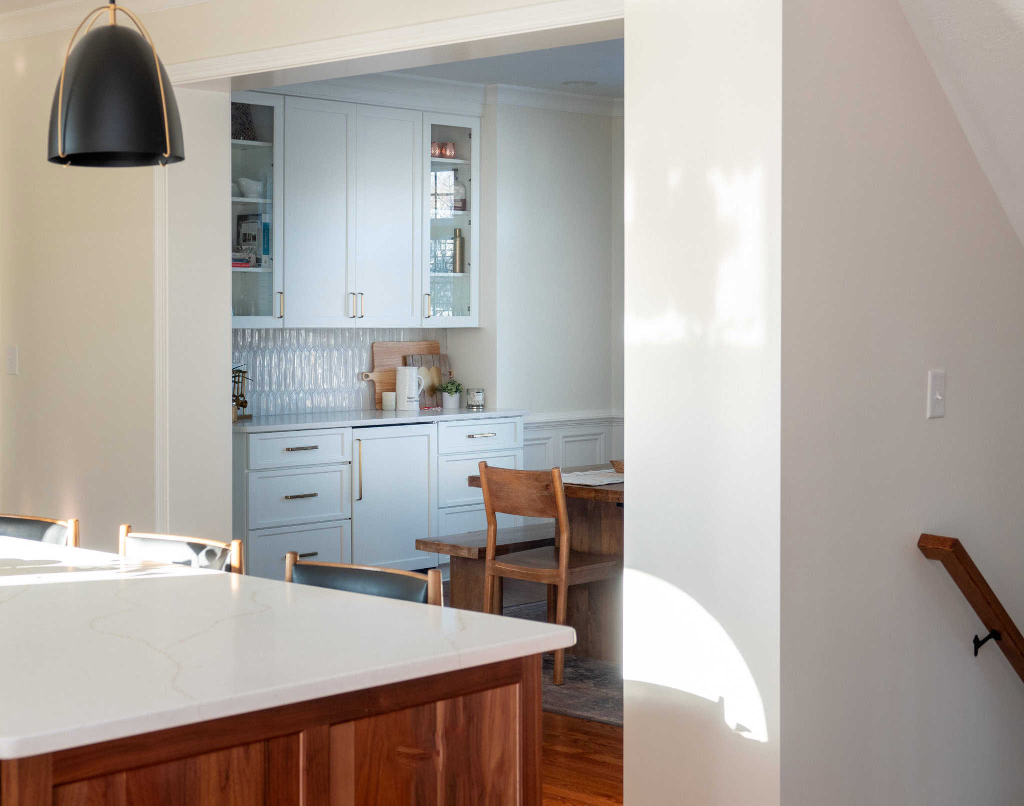 Nicholas Design Build | Kitchen with light gray cabinets, a wooden dining table, and chairs. Modern lighting fixture hangs over a large white countertop. Subtle natural light casts shadows on the walls.