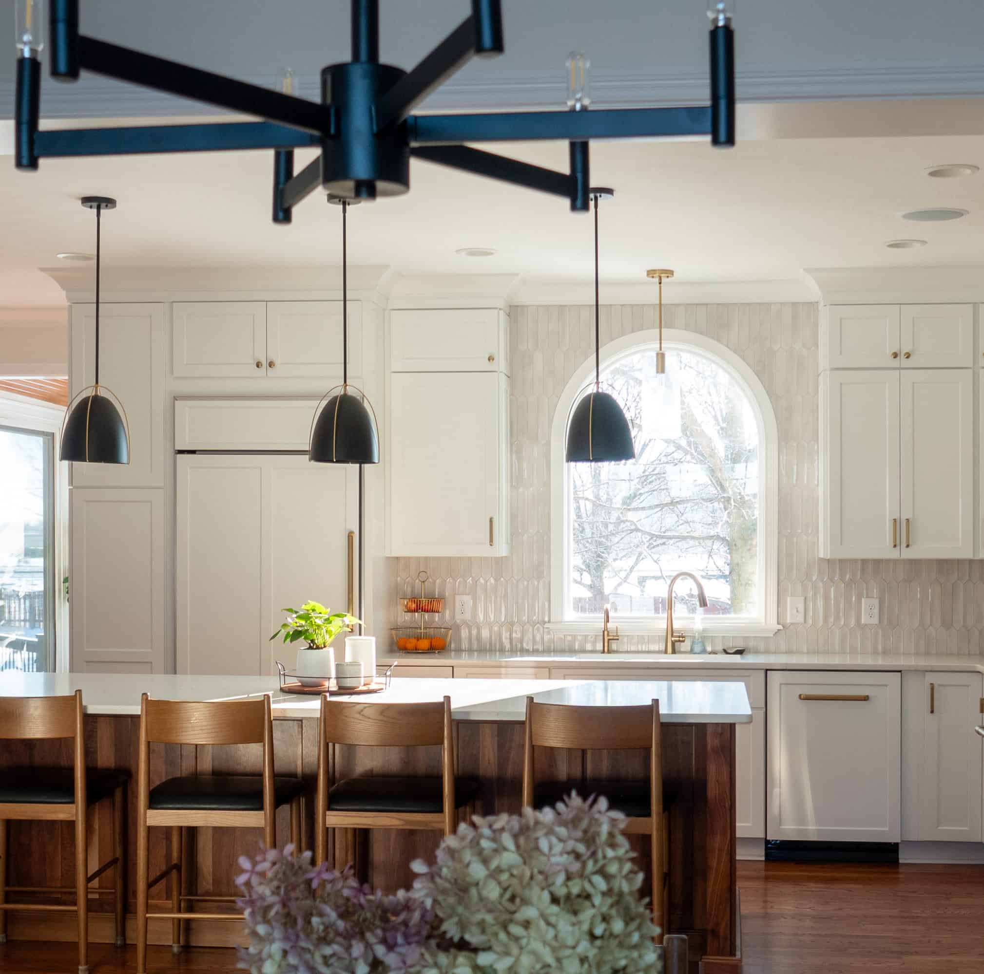 Nicholas Design Build | Modern kitchen with a large island, wooden stools, and pendant lights. White cabinets and an arched window offer natural light. A fruit bowl and plant decorate the counter.