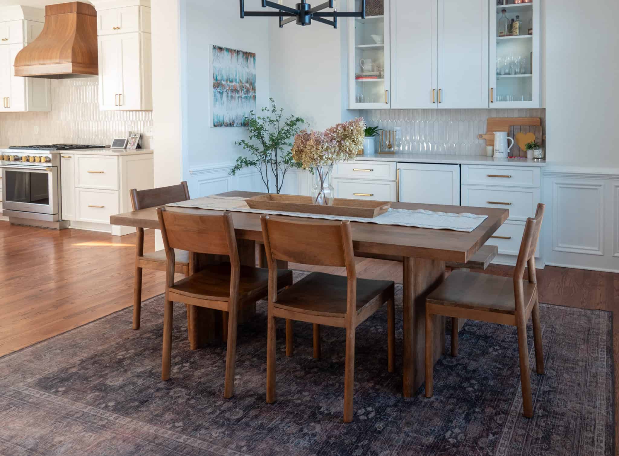 Nicholas Design Build | A dining room with a wooden table and six chairs on a dark patterned rug. White cabinets and a stove are in the background.